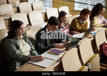 Group of multiethnic young students sitting in lecture hall taking notes and using digital devices during university class, focusing on studying and academic work Stock Photo