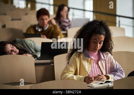 Young adult Black woman writing in notebook while sitting in lecture hall, multiethnic group of university students in background using laptops and resting during class Stock Photo