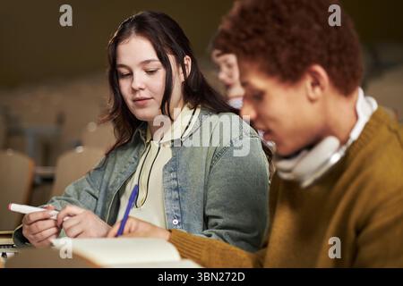 Caucasian young woman and Black young man studying together in university classroom, writing notes and discussing assignment, both focused on academic work, blurred student in background Stock Photo