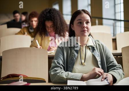 Happy Caucasian young woman sitting in university lecture hall holding open book, looking forward, multiethnic group of young students studying and writing in background Stock Photo