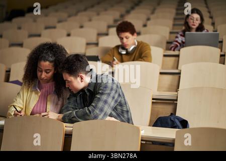 Multiethnic group of young university students sitting in lecture hall, two students collaborating on assignment in foreground while others working independently on laptops in background Stock Photo
