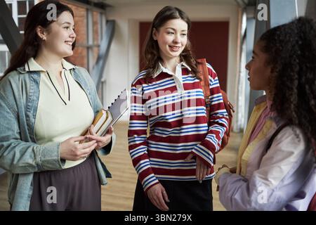 Three young women, including Caucasian and Black individuals, standing in university hallway, chatting and holding notebooks, engaging in conversation between classes Stock Photo