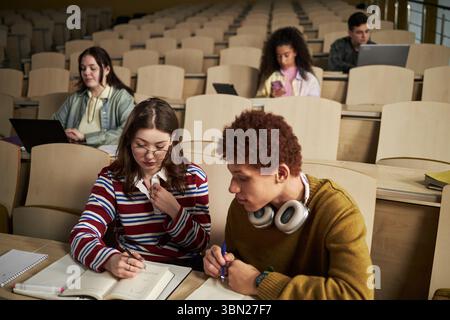 Group of multiethnic university students studying in lecture hall, young Caucasian woman and Black man collaborating on assignment while others using laptops and smartphone in background Stock Photo