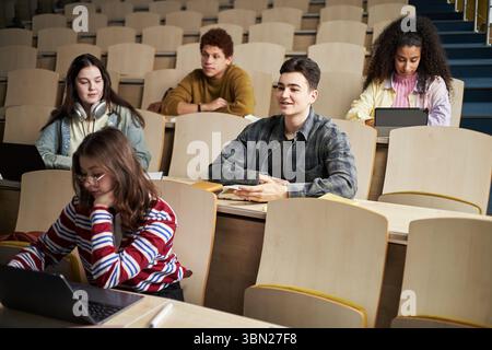 Group of multiethnic university students sitting in lecture hall listening to class, young Caucasian man smiling, busy woman using laptop, African American friends studying Stock Photo