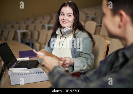 Caucasian young woman with long brown hair sitting in lecture hall receiving notebook from young man, wearing headphones around neck, studying with laptop and textbooks on desk Stock Photo