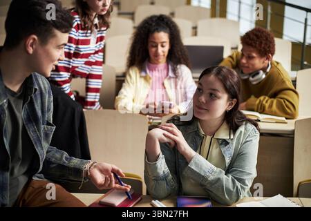 Group of multiethnic university students interacting in classroom, young Caucasian woman listening to male classmate while other students using smartphones and notebooks Stock Photo