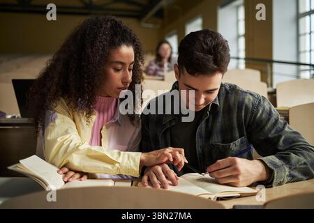 Young Caucasian man and Black woman studying together in university lecture hall, reading textbooks and taking notes, collaborating on academic assignment, focused on learning Stock Photo