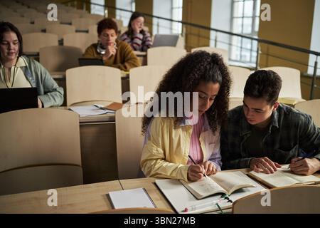 Group of multiethnic university students sitting in lecture hall, young adults collaborating on assignments, writing in notebooks, using laptops, studying together during class Stock Photo