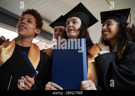 Diverse group of young university students wearing graduation caps and gowns standing together smiling and holding diploma folders during graduation ceremony Stock Photo