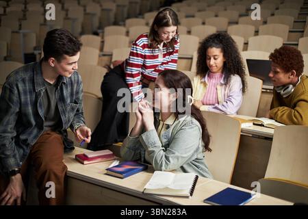 Group of multiethnic university students casually chatting in lecture hall,discussing notes and studying together with notebooks and textbooks on desks Stock Photo