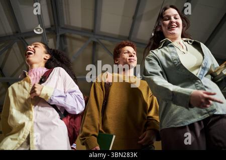 Group of diverse university students walking together indoors, multiethnic teenagers carrying backpacks and books, smiling and talking while moving through campus building Stock Photo