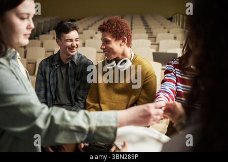 Group of multiethnic university students sitting in lecture hall interacting and smiling, young men and women engaging in conversation and teamwork, headphones visible around neck Stock Photo