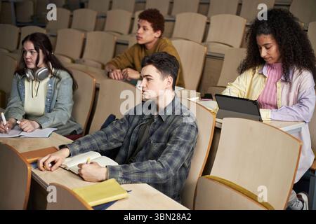 Group of multiethnic university students sitting in lecture hall taking notes and using digital tablet, listening to professor explaining new material, taking notes Stock Photo