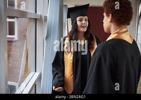 Caucasian young adlt woman and Black man wearing graduation caps and gowns standing indoors, talking near window, university students celebrating academic achievement Stock Photo