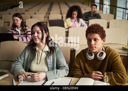 Group of multiethnic university students sitting in lecture hall listening to lesson, two teenagers in foreground taking notes with notebooks and laptops, others studying in background Stock Photo