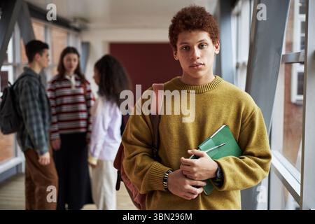 Portrait of young Black man standing in university hallway holding notebook and backpack, looking into camera while group of multiethnic students talking in background Stock Photo
