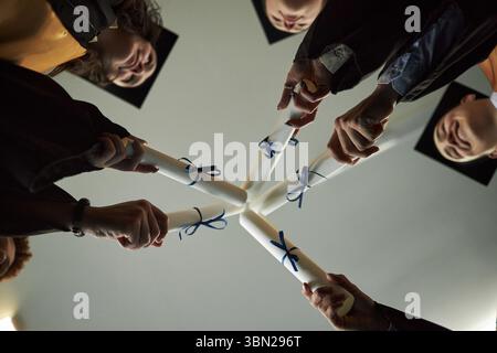 Group of diverse young university students wearing graduation caps holding diplomas together in circle celebrating academic achievement viewed from below Stock Photo