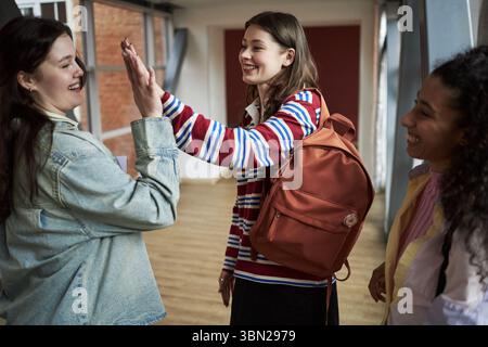 Group of multiethnic young adult women standing in university hallway smiling and giving high five, wearing backpacks and engaging in friendly conversation between classes on college campus Stock Photo