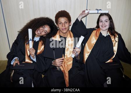 Diverse group of cheerful young students wearing graduation gowns sitting together smiling and holding diplomas, two women and man celebrating academic achievement at university Stock Photo