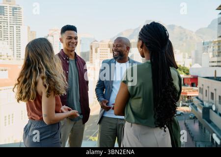 A diverse group of smiling business professionals talking on rooftop in city on sunny day Stock Photo