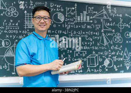 Portrait of Asian male teacher holding an open book and chalk in front of a chalkboard filled with mathematical formulas. Ideal for content about educ Stock Photo