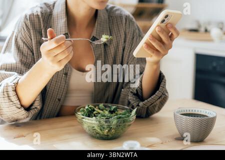 Unrecognizable modern woman having breakfast with salad and scrolling the news feed in her phone Stock Photo