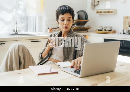Young woman sitting at home, drinking coffee and working on laptop Stock Photo