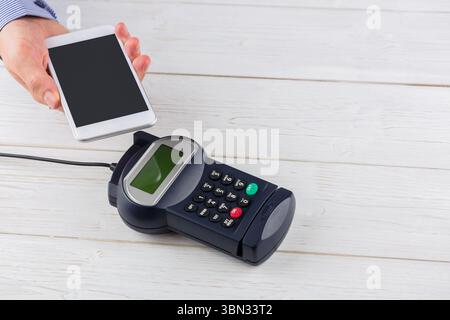 Man is holding smartphone above POS terminal with cable on pale wooden tabletop in flat design Stock Photo