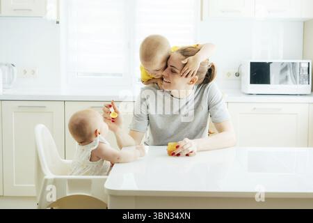 Family fun, mom feeds the children in the kitchen. Mom feeds the baby with false vegetable puree, the older child hugs mom. Family pleasure moments on Stock Photo