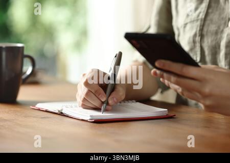 Close up portrait of a woman hands writing in paper agenda checking smart phone on wooden table at home Stock Photo