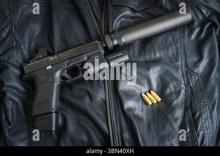 A tactical pistol with a silencer, red dot sight, and flashlight lies on a black leather jacket next to ammunition. Stock Photo