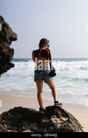 Woman traveler with camera taking pictures of ocean from rock on beach Stock Photo