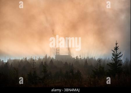 Sun rises, its rays pierce through the mist, casting dramatic beams of light over landscape. Antenna tower and building emerge from fog, surrounded by dense forest of pine trees. Stock Photo