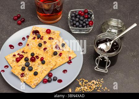 Biscuits with berries on gray plate. Sunflower seeds and flaxseed on table. Jar of jam, glass of tea and blueberries in bowl. Black background. Top vi Stock Photo