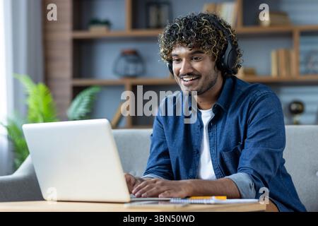 A smiling man with headphones uses a laptop while working or attending a video call indoors. Stock Photo