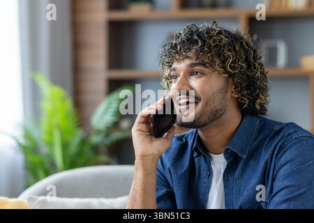 A smiling man with curly hair is talking on a phone indoors, appearing relaxed. Stock Photo
