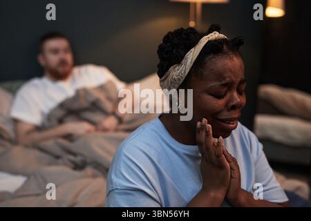 Black woman crying with hands clasped near face, sitting on bed foreground while Caucasian man with beard sitting in background under blanket looking away during conflict Stock Photo