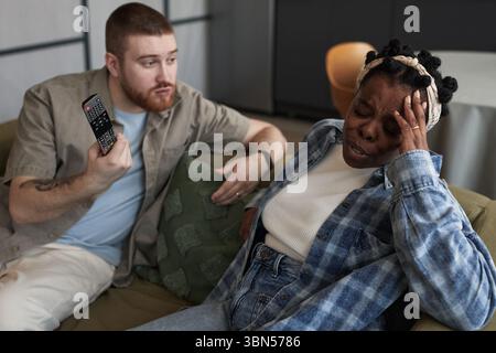 Middle aged Caucasian man holding remote control gesturing while middle aged Black woman sitting beside him touching forehead appearing upset during conflict on couch Stock Photo