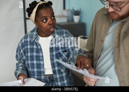 Black young adult woman confronting Caucasian young adult man while holding documents, both standing close together, woman gesturing emphatically and man looking down at papers Stock Photo