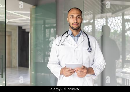A doctor, wearing a stethoscope and lab coat, stands in an office holding a tablet. He appears calm and professional. Stock Photo