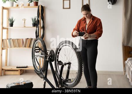 Young woman standing next to her bicycle at home taking a photo of it with her smartphone. DIY concept. Capturing details for reference or sharing. Stock Photo