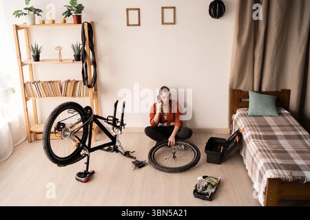 Female student repairing bicycle at home holding bike wheel in hands calling support hotline. Bike maintenance and repairing concept. High angle view Stock Photo