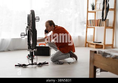Man fixing bike indoors and speaking on phone with technical support for repair advice. Concept of home maintenance, bicycle repair, and remote mechan Stock Photo