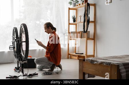 small business of mechanic. Young man repairing bicycle doing maintenance of bike using mobile phone at home. Copy space Stock Photo