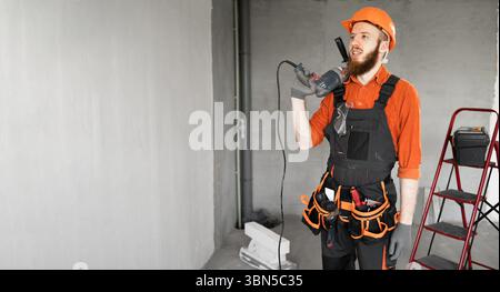 Bearded male construction worker or builder holding electric drill or perforator standing in new house. profession, construction and building concept. Stock Photo