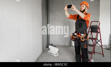 Young bearded construction worker in helmet and tool belt taking picture of wall for report using cellphone at construction site Stock Photo