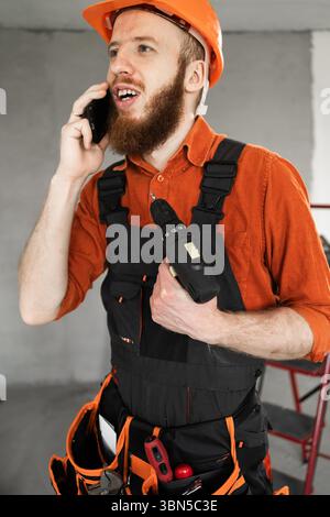 Foreman calls on phone. Builder calling using phone. Bearded man wears construction workers uniform. Repairman in helmet. Copy space Stock Photo