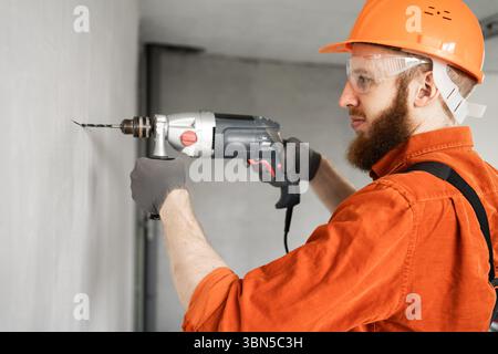 Portrait of concentrated bearded construction worker drilling hole with electric drill in wall. Professional builder and foreman concept Stock Photo