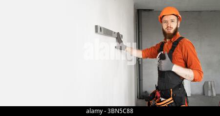 Repair and construction concept. male builder or civil engineer in protective helmet measures wall with level. Construction worker checking wall. Bann Stock Photo