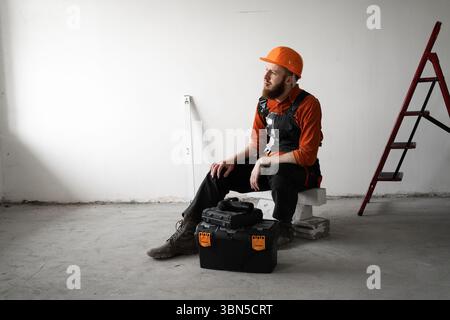Builder or engineer in helmet and blue overalls sitting on construction site near toolbox looking thoughtfully into distance. Copy space Stock Photo